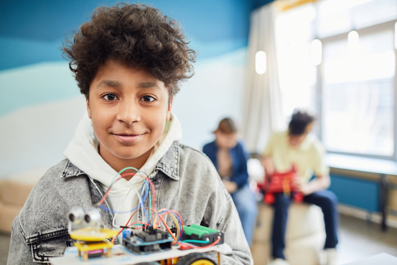 A young boy smiling confidently while holding a DIY robotics project in a classroom setting.